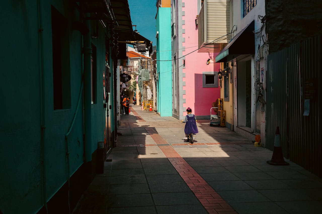 A child on a scooter explores a vibrant alleyway in Macau, filled with colorful buildings and warm sunlight.